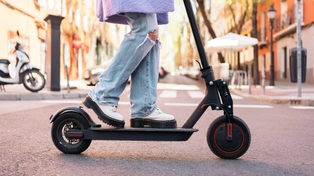 Persona montando patinete negro en calle urbana, con edificios y una moto al fondo.
