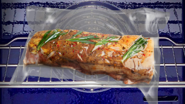 Un horno azul contiene una bolsa de plástico transparente con un trozo de carne sazonada en una rejilla metálica.