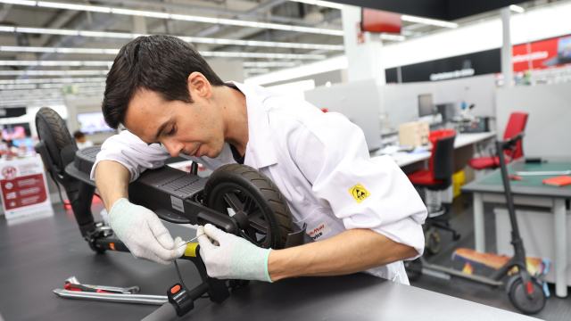Un hombre con guantes blancos y bata examina un patinete negro sobre una mesa. En el fondo, se ven más patinetes y mobiliario de oficina.