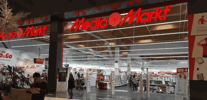 Entrada a tienda Media Markt con letrero rojo, gente comprando y estanterías con productos.