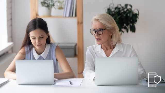 Dos mujeres en un escritorio blanco, ambas usando laptops. La mujer a la derecha tiene gafas y una camisa blanca.