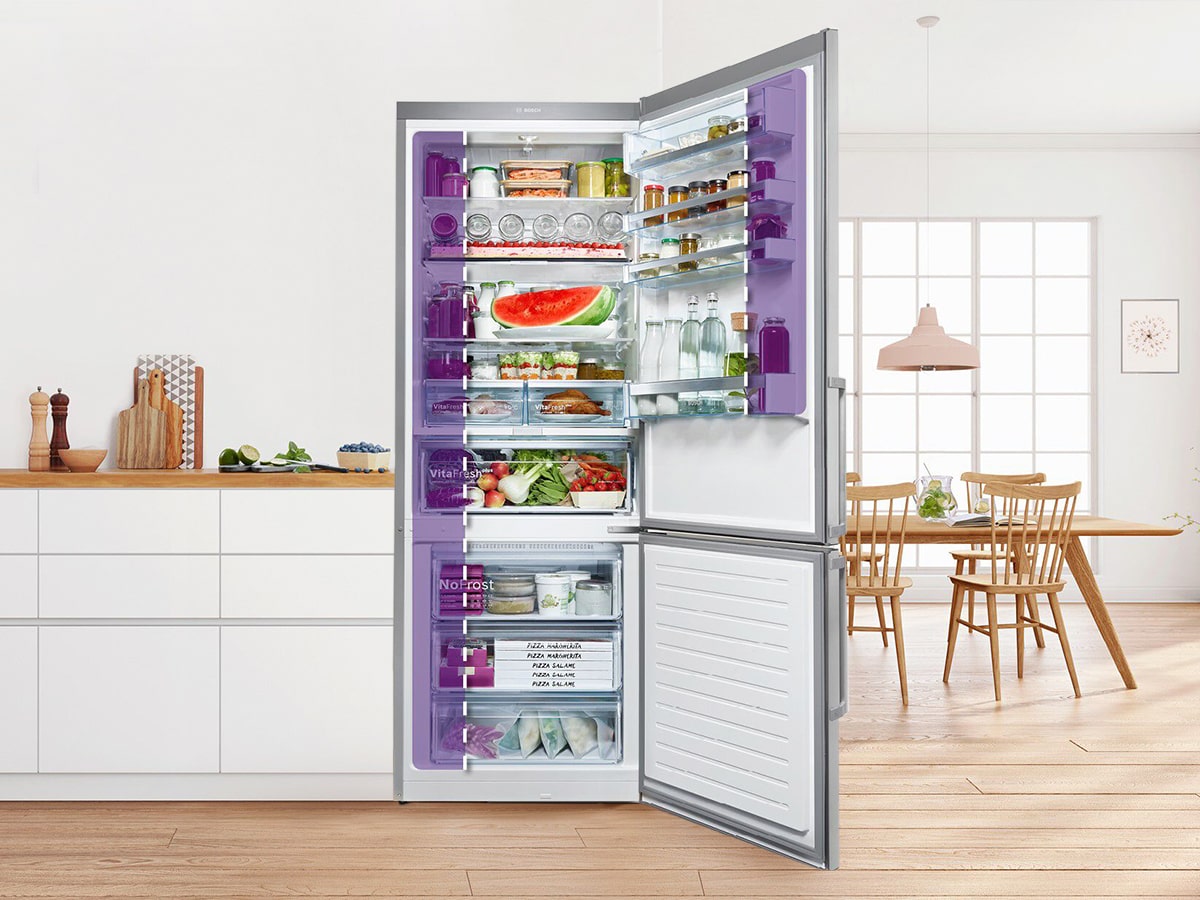 Un refrigerador abierto muestra alimentos en estantes y cajones. Se ve una cocina con mesa, sillas y encimera blanca.