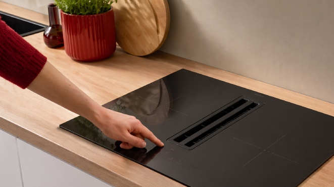 Placa de inducción negra con extractor central, mano tocando controles, encimera de madera y detalles rojos en cocina moderna.