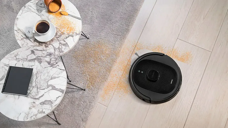 Vista elevada de una aspiradora robot negra cerca de mesas con café y un cuaderno sobre una alfombra gris y un suelo de madera.