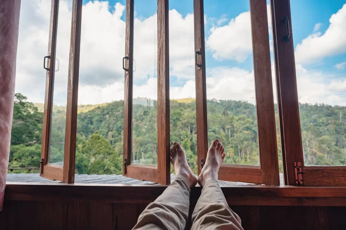 `Persona descansando con las piernas estiradas junto a una ventana abierta de madera, con vistas a un paisaje de montaña y bosque bajo un cielo azul con nubes.`