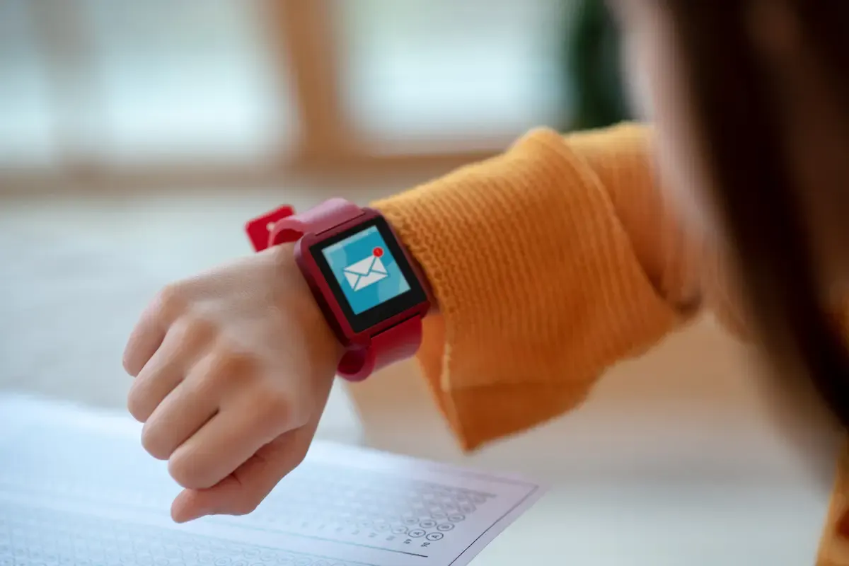 Mano con reloj rojo mostrando un icono de correo. La persona viste una camisa naranja y hay un papel con círculos en la mesa.