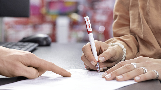 Una persona firma un documento con un bolígrafo blanco y rojo mientras otra persona señala el papel. Hay un ordenador al fondo.