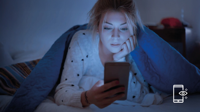 Una persona en cama mira un teléfono móvil bajo una manta azul, con una luz tenue y un icono de filtro de luz azul.
