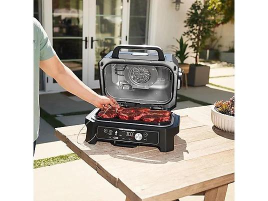 Una persona cocina carne en una parrilla eléctrica negra sobre una mesa de madera al aire libre. Hay plantas y una casa cerca.