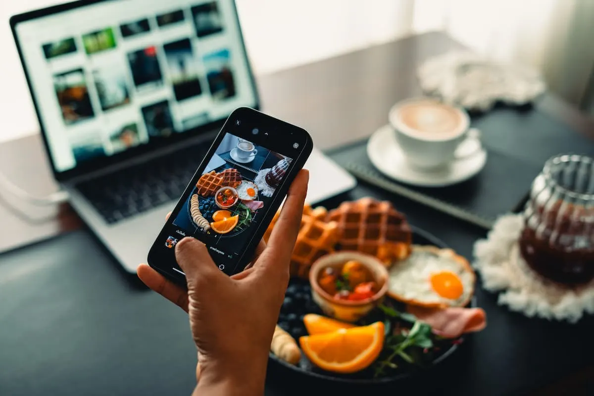Persona fotografiando con su móvil un desayuno con gofres, huevos y fruta, junto a un portátil y una taza de café.