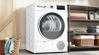 Secadora con bomba de calor, instalada en un amplio cuarto de lavado, junto a una estantería con ropa limpia y un cesto de ropa para lavar