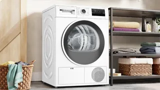 Secadora con bomba de calor, instalada en un amplio cuarto de lavado, junto a una estantería con ropa limpia y un cesto de ropa para lavar