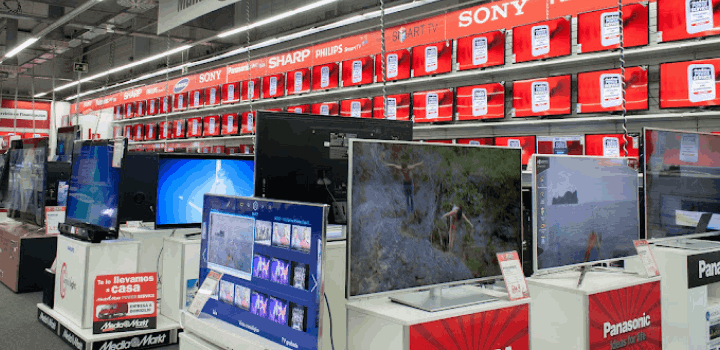 Televisores de varias marcas como Sony, Sharp y Panasonic en una tienda, exhibidos sobre bases blancas y con estantes rojos.