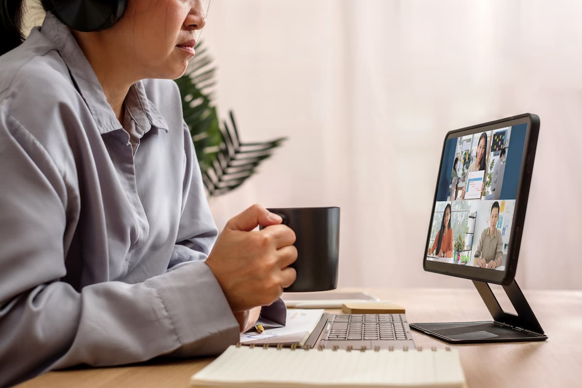 Persona en videollamada frente a una tablet sobre soporte en un escritorio con teclado y cuaderno.