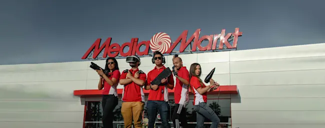 Cinco personas con camisetas rojas posan frente a un edificio de Media Markt. Sostienen accesorios electrónicos y de juego.