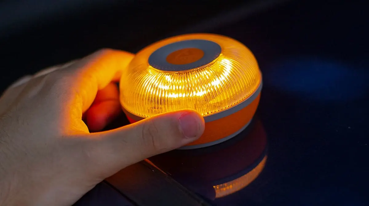 Mano sujetando una baliza naranja y gris. La baliza emite una luz amarilla brillante y está sobre una superficie oscura y reflectante.