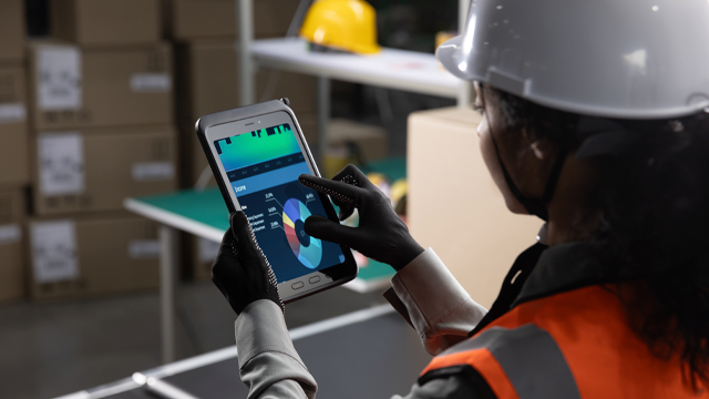 Trabajador con chaleco naranja y casco blanco usa tableta con gráfico circular en almacén con cajas y estantería.