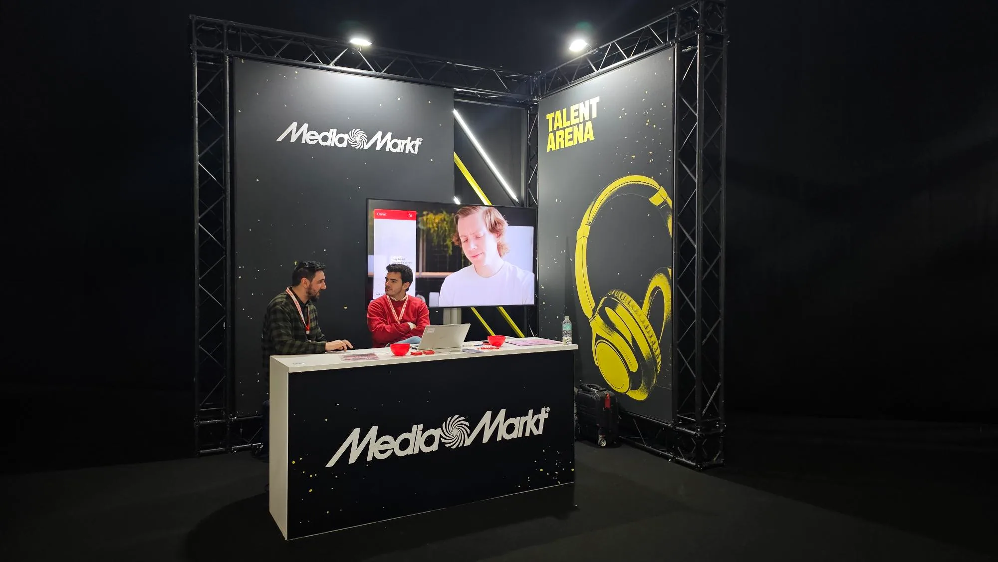 Dos hombres en un stand de Media Markt con un monitor que muestra a una persona y auriculares amarillos.