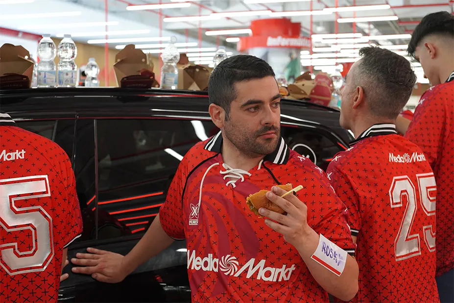 Un grupo de personas con camisetas rojas están reunidas alrededor de un coche negro, comiendo y charlando.
