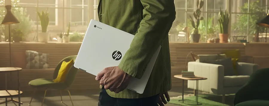 Un hombre con camisa verde lleva un portátil HP blanco en una sala de estar con plantas y muebles modernos.