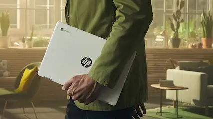 Un hombre con camisa verde lleva un portátil HP blanco en una sala de estar con plantas y muebles modernos.