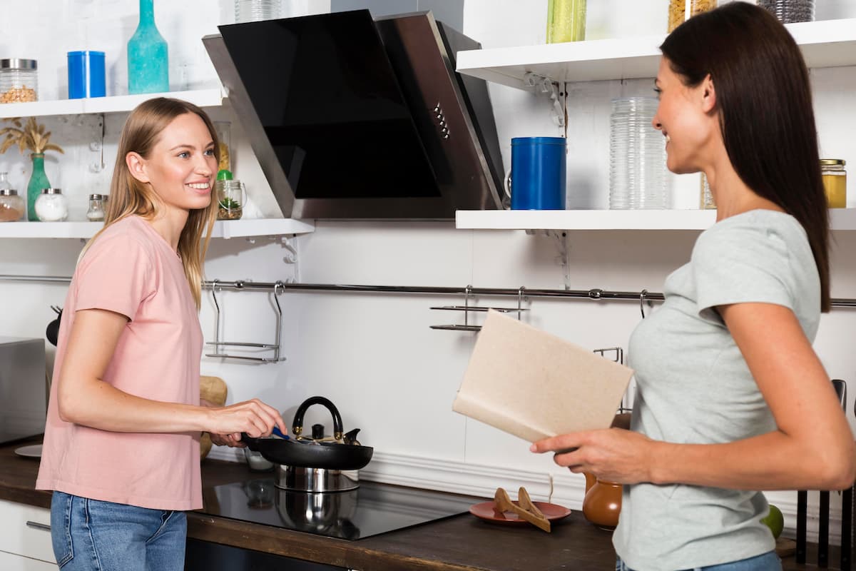 Dos mujeres charlan en una cocina moderna mientras una cocina en la sartén y la otra sostiene un libro de recetas.