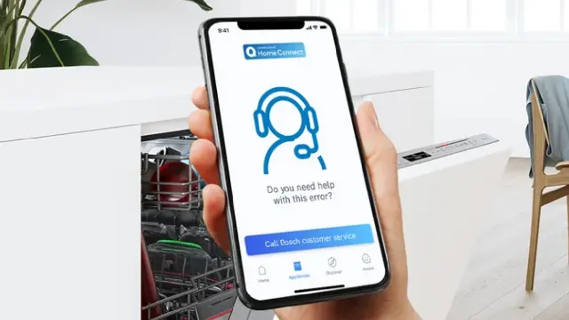 Mano sosteniendo teléfono con el servicio de Asistencia Remota de la Aplicación Home Connect. Cocina con lavavajillas abierto en el fondo.