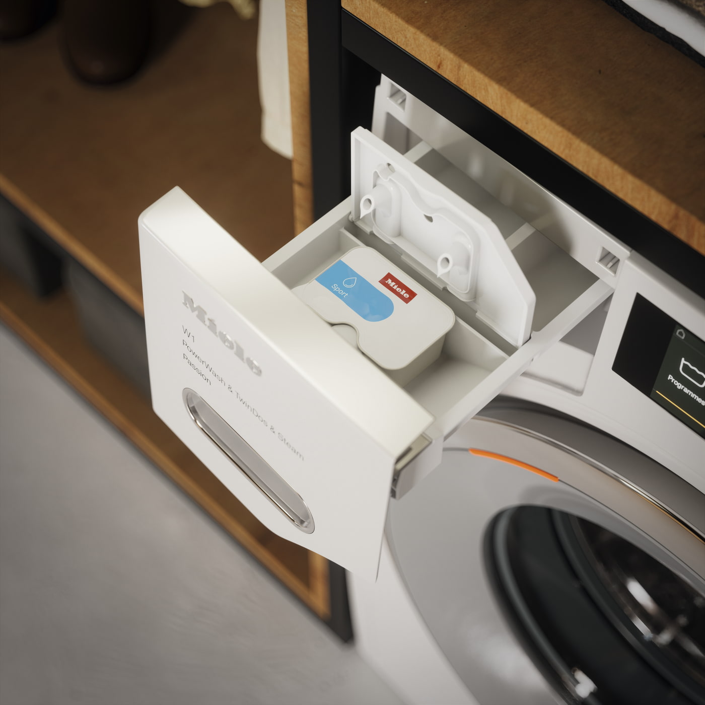 Lavadora Miele con cajón dispensador abierto, mostrando compartimento para detergente deportivo. Estantes de madera visibles arriba y a la izquierda.