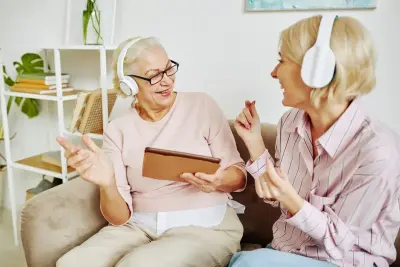 Dos mujeres con auriculares están sentadas en un sofá. Una sostiene una tableta marrón. Hay estanterías y plantas en el fondo.