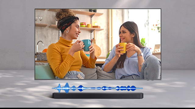 Dos mujeres conversan y toman café en una sala, vistas en la pantalla de un televisor con barra de sonido y ondas de sonido azules.