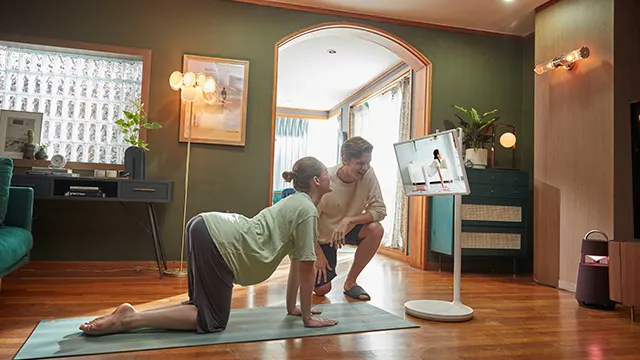Una mujer hace yoga en una esterilla mientras un hombre observa una pantalla en una habitación.