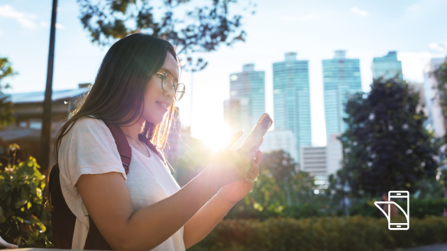 Una persona usa un teléfono en un parque urbano con edificios altos al fondo y árboles y plantas alrededor.