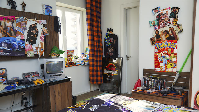 Habitación con pósteres, videojuegos, una televisión y una cama con motivos de dibujos animados. Ventana con cortinas a cuadros.
