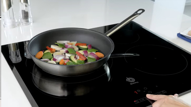 Detalle de una sartén en la que se cocinan distintas hortalizas y un dedo ajustando la potencia de la zona de cocción.