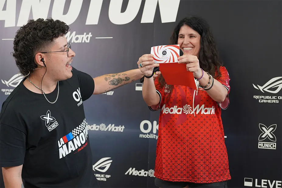Dos personas sonriendo frente a un panel con logos. Una mujer sostiene un sobre rojo y blanco. Un hombre con audífonos mira el sobre.