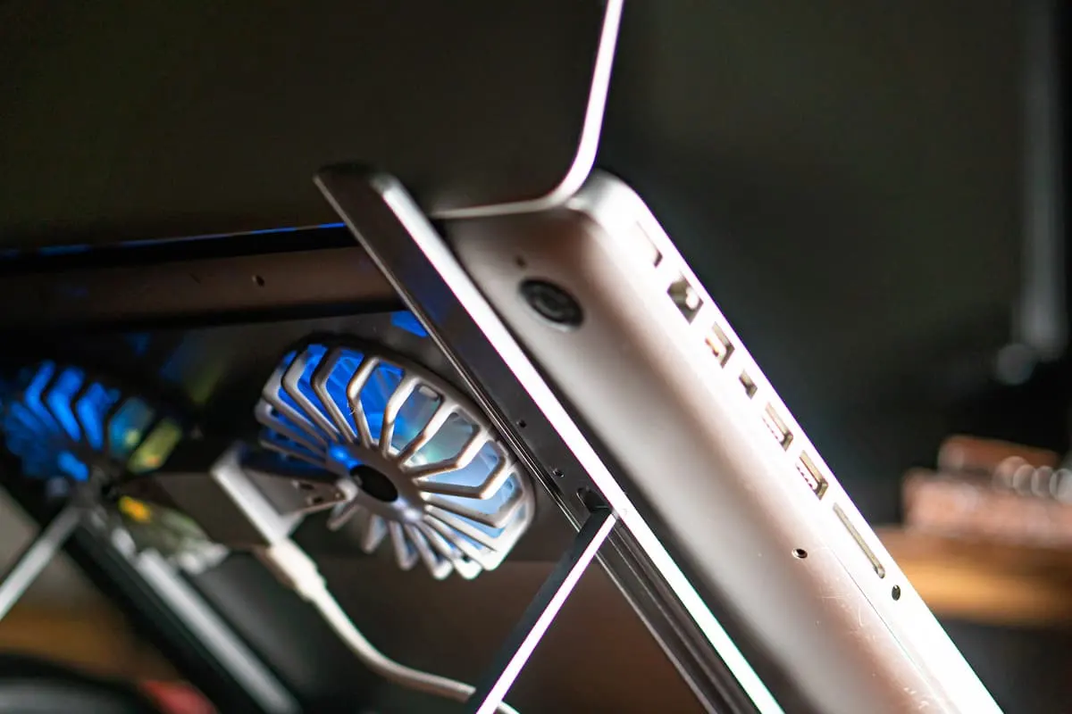 Portátil plateado visto de perfil sobre una base inclinada con ventilador de refrigeración USB iluminado en azul, mostrando los puertos laterales del equipo.