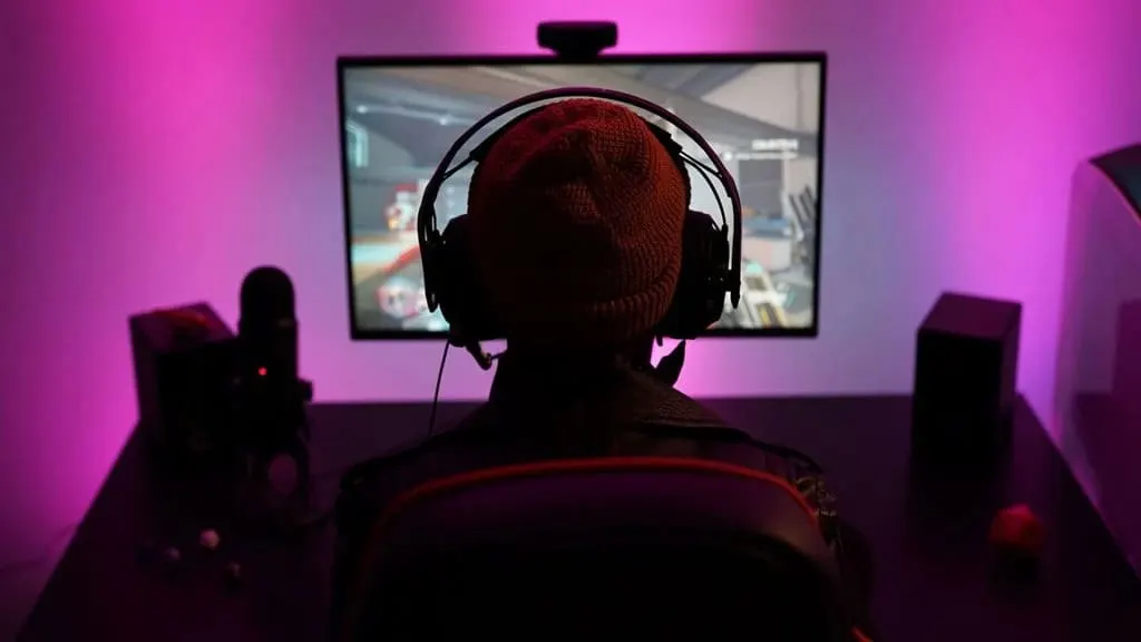Un individuo con gorro y auriculares frente a un monitor que muestra un videojuego, iluminado con luz morada. Hay altavoces y un micrófono en la mesa.
