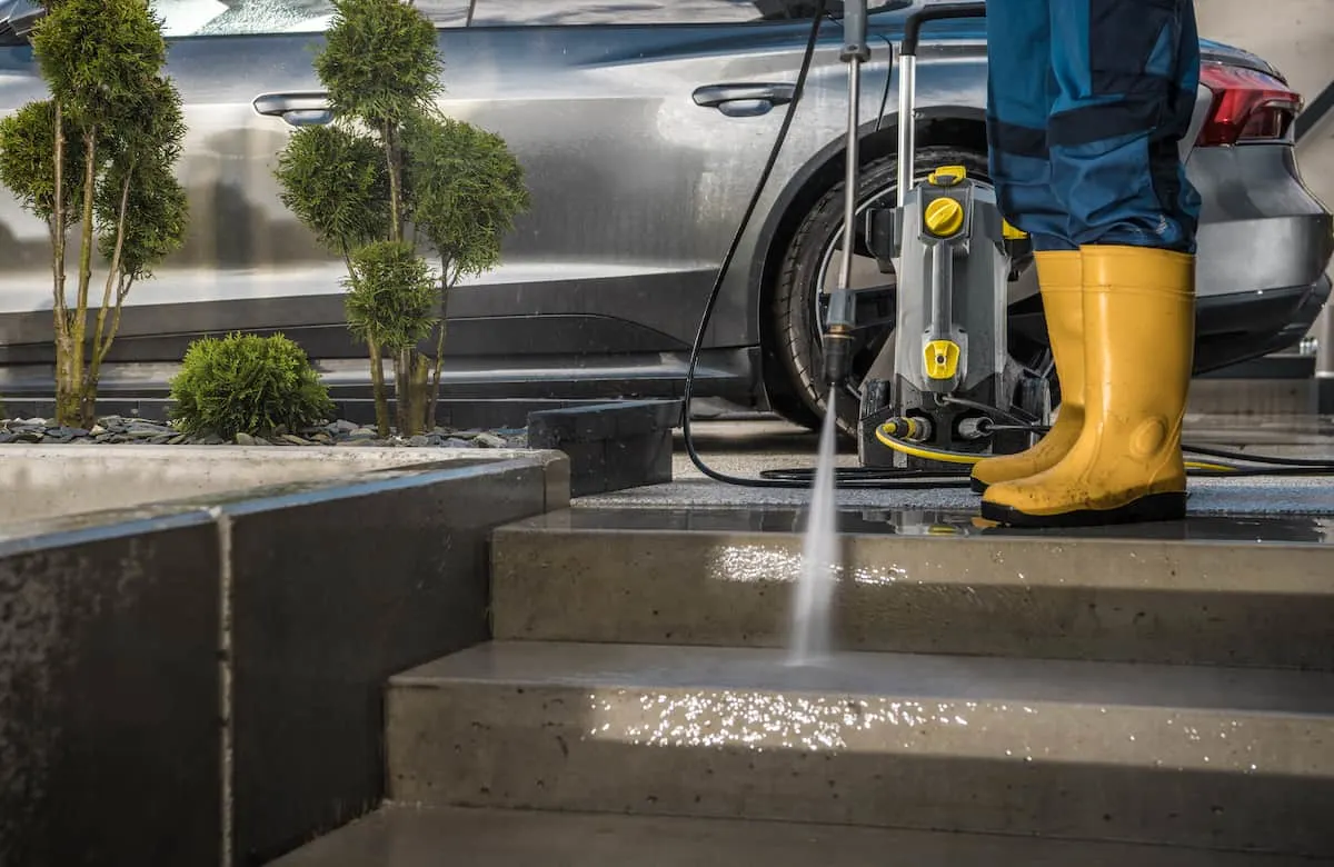 Persona con botas amarillas usando una hidrolavadora a presión para limpiar escalones de concreto, con un auto y plantas al fondo.