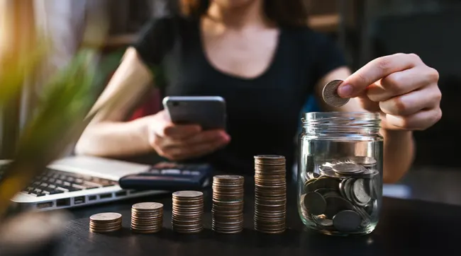 Una persona mete una moneda en un tarro lleno de monedas. Hay pilas de monedas, un móvil y un ordenador portátil.