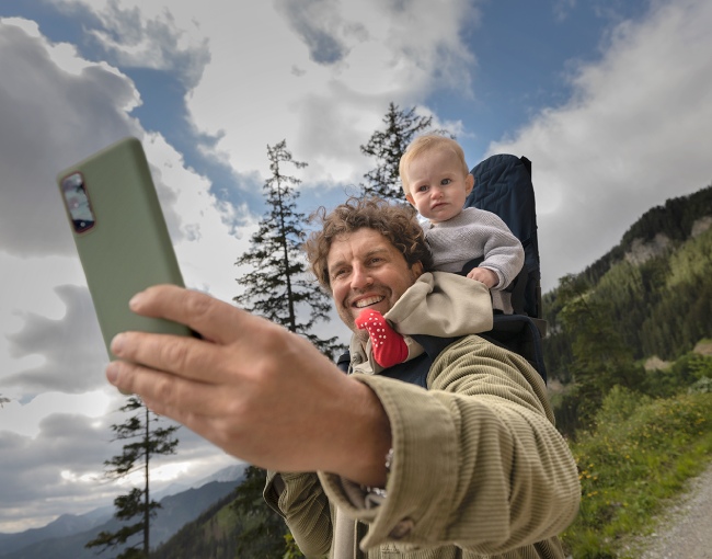 Padre sonriente toma selfie con un teléfono verde; el bebé está sentado en un portabebés. El fondo tiene árboles y cielo nublado.