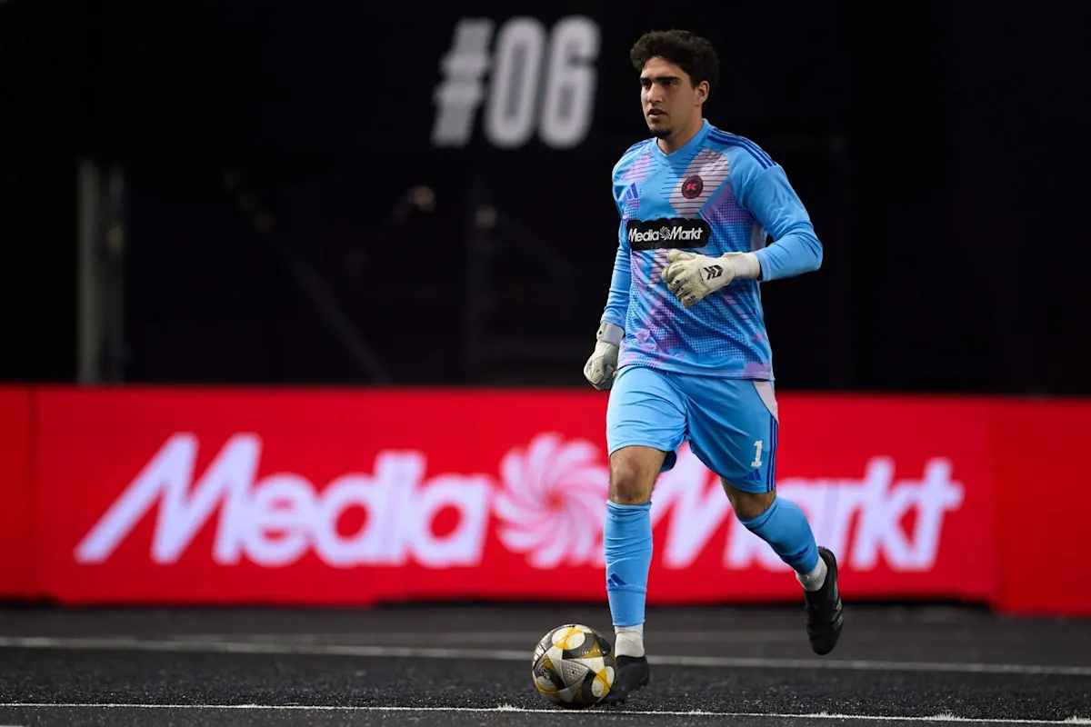 Un jugador de fútbol con uniforme celeste y guantes blancos controla un balón frente a un anuncio rojo de "MediaMarkt".