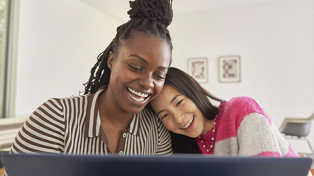 Dos mujeres sonriendo frente a un ordenador portátil; una lleva un suéter rosa y blanco, la otra una camisa de rayas marrones y blancas.