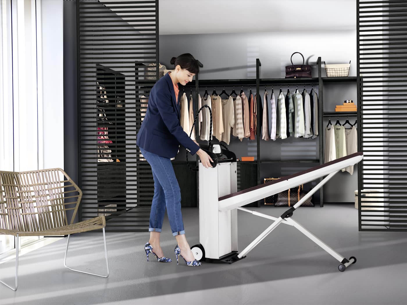 Mujer con blazer azul y jeans junto a plancha vertical con tabla inclinada. Al fondo, perchero y silla de mimbre.