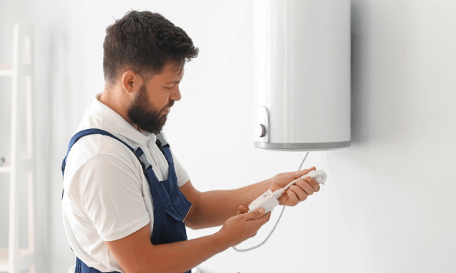 Un hombre con overol azul sostiene un enchufe blanco cerca de un calentador de agua blanco montado en una pared blanca.