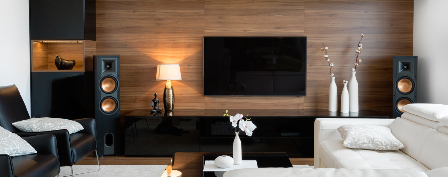 Salón con pared de madera, TV, altavoces negros, lámpara, sillón y sofá blancos. Hay jarrones y adornos decorativos.