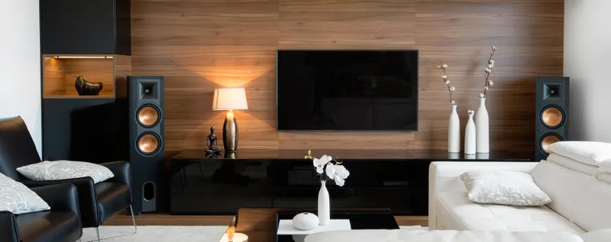 Salón con pared de madera, TV, altavoces negros, lámpara, sillón y sofá blancos. Hay jarrones y adornos decorativos.