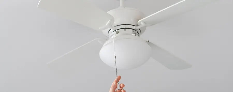 Un ventilador blanco de cinco aspas está montado en el techo. Una mano tira de la cadena del ventilador.