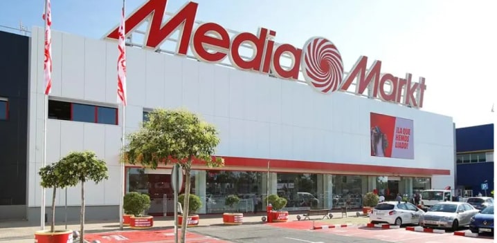 Edificio blanco de Media Markt con letras rojas, banderas y árboles en macetas rojas. Hay coches aparcados.
