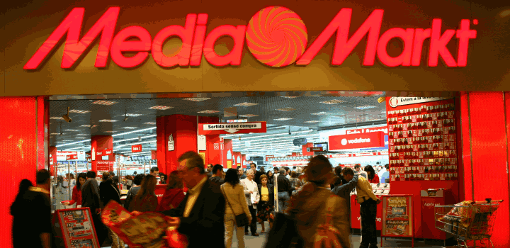 Entrada a tienda con letrero rojo "Media Markt" y multitud de personas entrando y saliendo; estanterías y anuncios visibles.