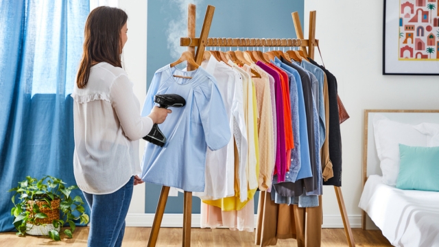 Una persona sostiene un vaporizador de ropa blanco y negro con detalles naranjas y su base, sobre un fondo rojo.